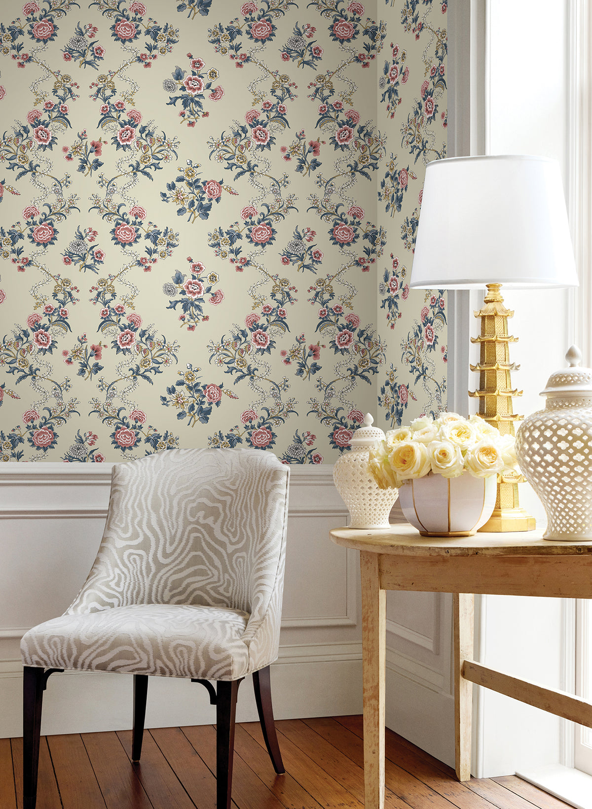 A white chair sits beside a wooden table topped with a lamp, a ceramic jar, and a bouquet of white roses. The wall features York Wallcoverings’ Genevieve Floral Trail Wallpaper in pink, blue, and green tones, while sunlight streams in through a nearby window.