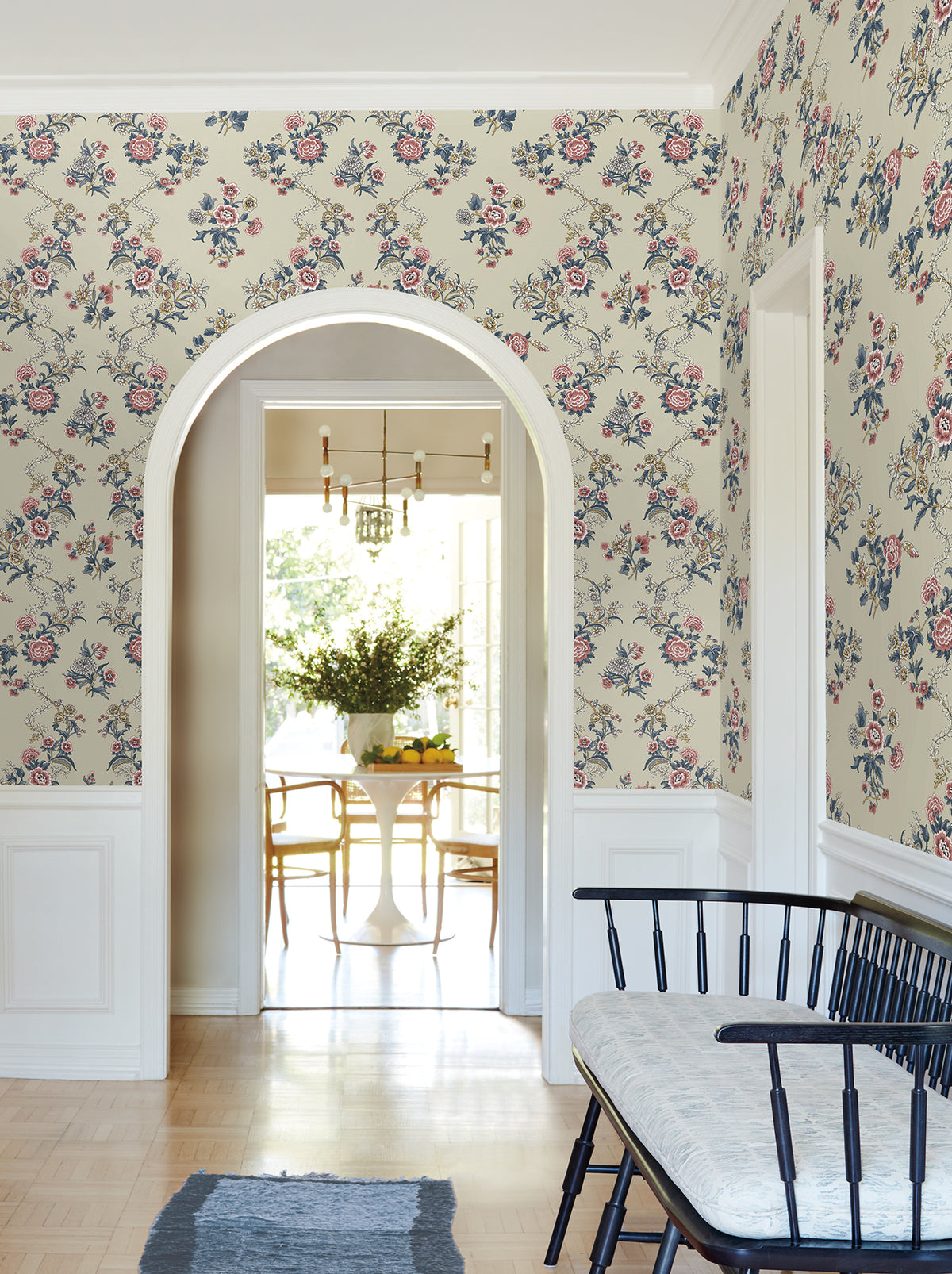 A hallway adorned with York Wallcoverings’ Genevieve Floral Trail Wallpaper features a black bench with a cushion and an archway that leads to a sunlit dining area, where a white table surrounded by wooden chairs is topped with a vase of greenery.