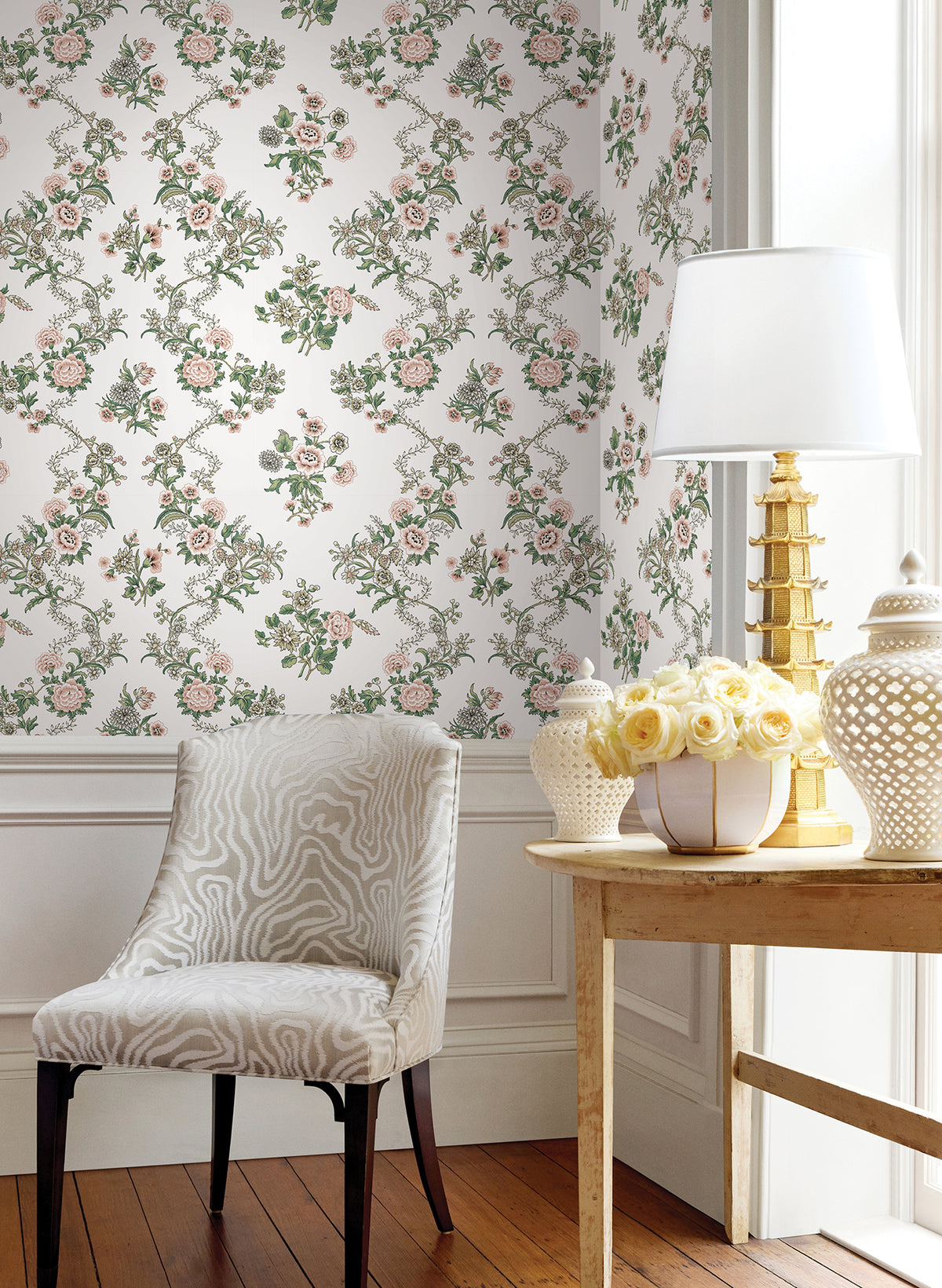 A white chair with a zebra pattern sits by a wooden table topped with a gold lamp, white vase of yellow roses, and a decorative jar. Genevieve Floral Trail Wallpaper by York Wallcoverings and sunlight brighten the elegant room.
