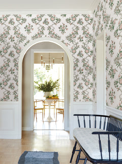 A hallway adorned with Genevieve Floral Trail Wallpaper by York Wallcoverings, featuring a black bench with a cushion, and an archway opening into a bright dining room with a white table, wooden chairs, and a vase of flowers.