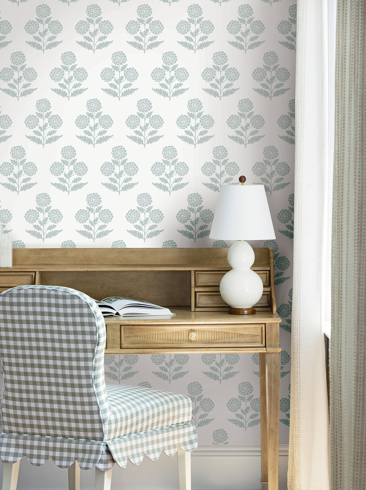 A wooden desk with a white table lamp, open book, and plaid upholstered chair is set against a wall adorned with York Wallcoverings’ Souci Marigolds wallpaper, next to a window with light curtains.