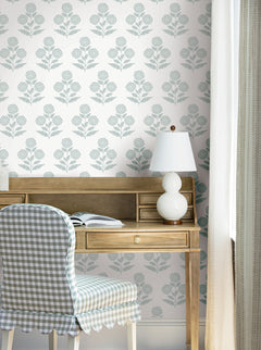 A wooden desk with a white table lamp, open book, and plaid upholstered chair is set against a wall adorned with York Wallcoverings’ Souci Marigolds wallpaper, next to a window with light curtains.
