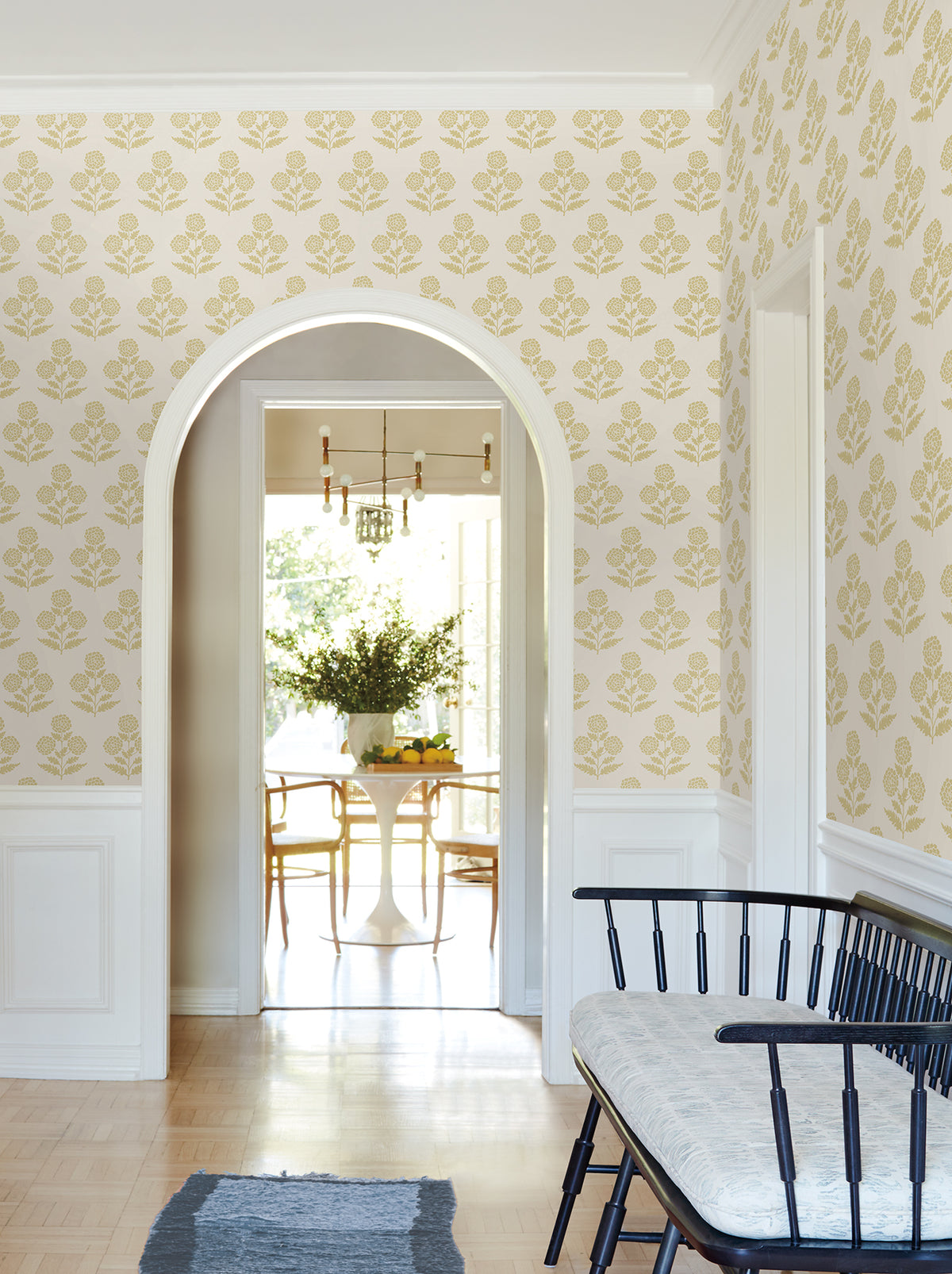 A hallway adorned with Souci Marigolds Wallpaper by York Wallcoverings features an archway that opens to a bright dining room with a round white table, wooden chairs, a black bench, and a rug atop light wood flooring.