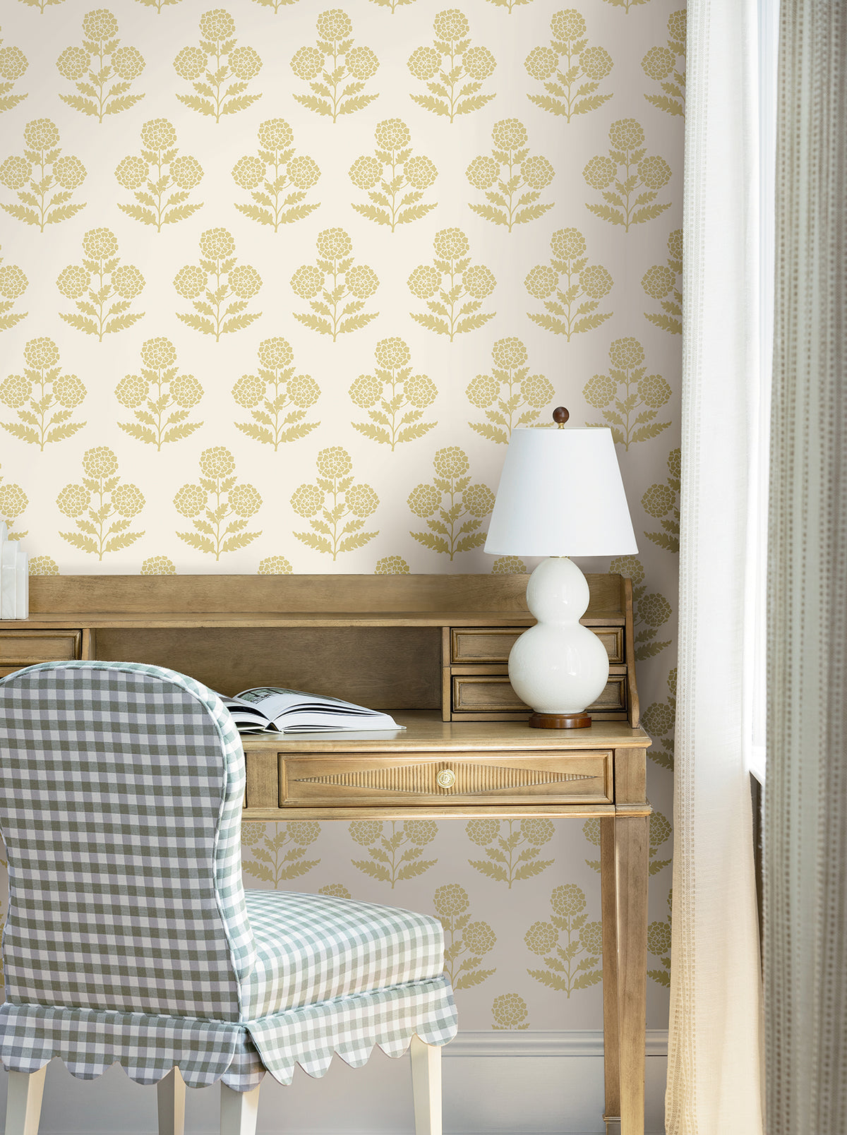 A wooden desk with an open book, a white lamp, and a blue gingham chair sits against a cream wall adorned with York Wallcoverings’ Souci Marigolds Wallpaper. Sunlight streams in through a window with light curtains.