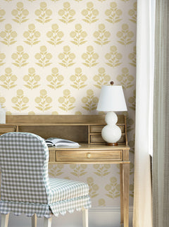 A wooden desk with an open book, a white lamp, and a blue gingham chair sits against a cream wall adorned with York Wallcoverings’ Souci Marigolds Wallpaper. Sunlight streams in through a window with light curtains.