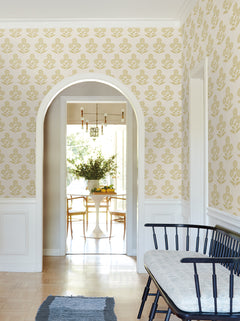 A hallway adorned with York Wallcoverings’ Souci Marigolds Wallpaper in yellow and accented by white trim leads to a bright dining room featuring a round table, chairs, and a vase of greenery. Along the hallway wall, a black bench with a cushion completes the scene.