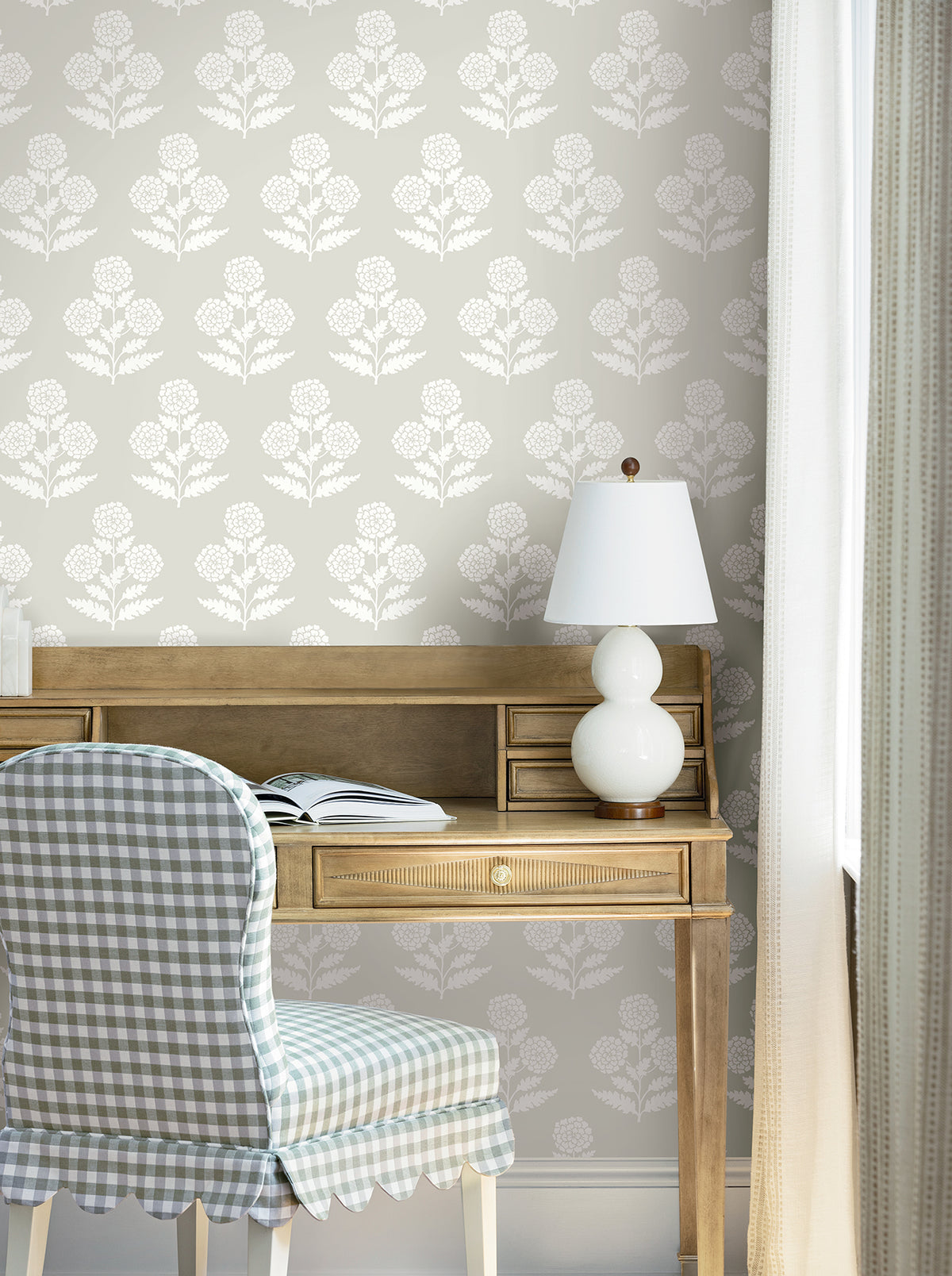 A wooden desk with an open book and white lamp sits against York Wallcoverings’ Souci Marigolds wallpaper. A chair with blue gingham fabric is in front of the desk, and light curtains hang to the right.