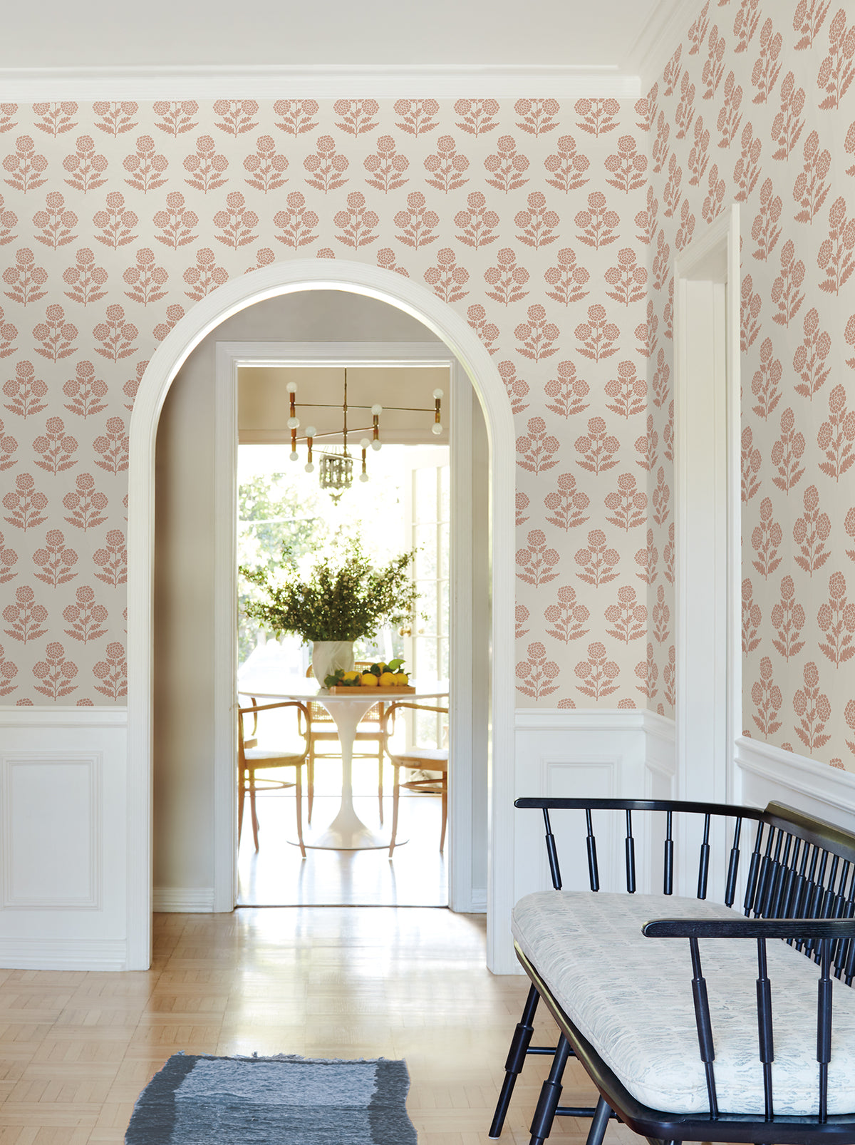 A hallway adorned with York Wallcoverings’ Souci Marigolds Wallpaper in pink, featuring a black bench with a cushion, a blue rug, and an arched doorway that opens into a sunlit dining area with a white table, chairs, and a flower arrangement.