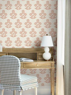 A wooden desk with an open book, a white lamp, and a checkered chair sits in front of a window. The wall behind is adorned with York Wallcoverings’ Souci Marigolds Wallpaper, featuring a pink floral pattern on a cream background. Light curtains frame the scene.
