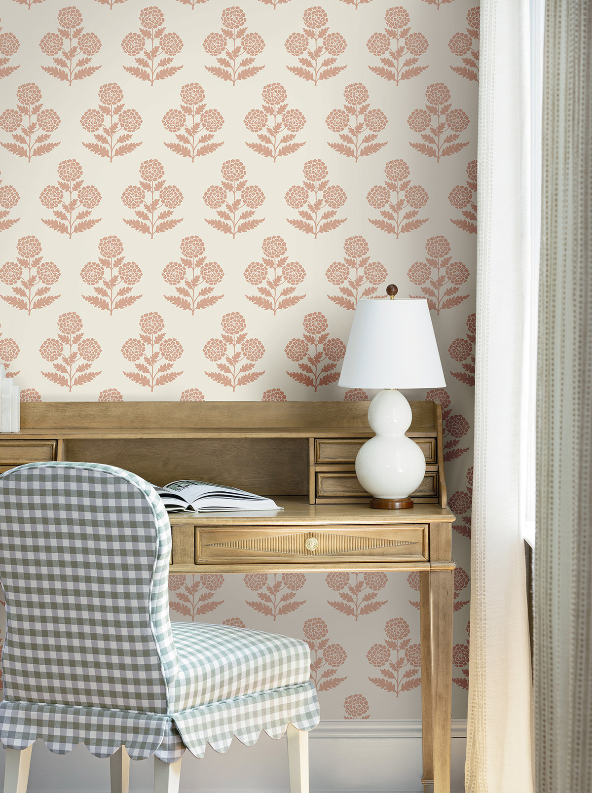 A wooden desk with an open book, white lamp, and a blue plaid chair sits in front of a window. The wall is adorned with York Wallcoverings’ Souci Marigolds Wallpaper. White curtains hang at the side of the window.