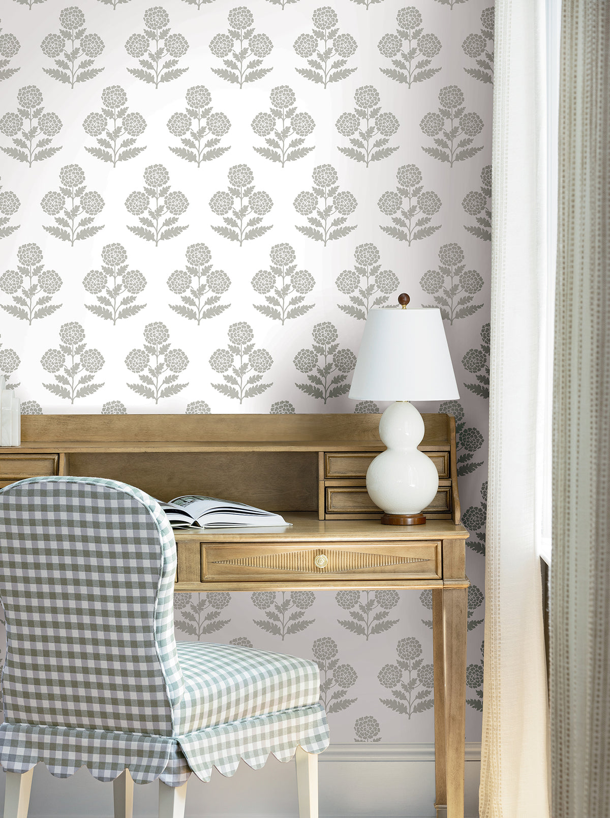 A wooden desk with an open book, white lamp, and checkered chair sits by a window with light curtains. The wall features the Souci Marigolds Wallpaper by York Wallcoverings in muted gray tones.