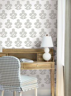 A wooden desk with an open book, a white lamp, and a chair with a blue checkered cover sits in front of a wall adorned with York Wallcoverings’ Souci Marigolds Wallpaper. Light curtains frame the scene.