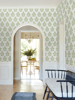 A hallway featuring Souci Marigolds Wallpaper by York Wallcoverings, an arched doorway, a black bench with a cushion, and a view into a dining area with a round table, chairs, chandelier, and vase of flowers.