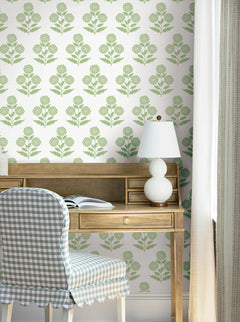 A wooden desk with an open book, a white lamp, and a checked chair sits in front of York Wallcoverings’ Souci Marigolds Wallpaper and airy sheer curtains, creating a cozy, light-filled workspace.