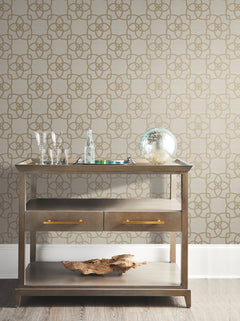 A small wooden bar cart with two drawers, glasses, a bottle, a decorative silver sphere, and a driftwood piece sits against Serendipity Wallpaper by York Wallcoverings.