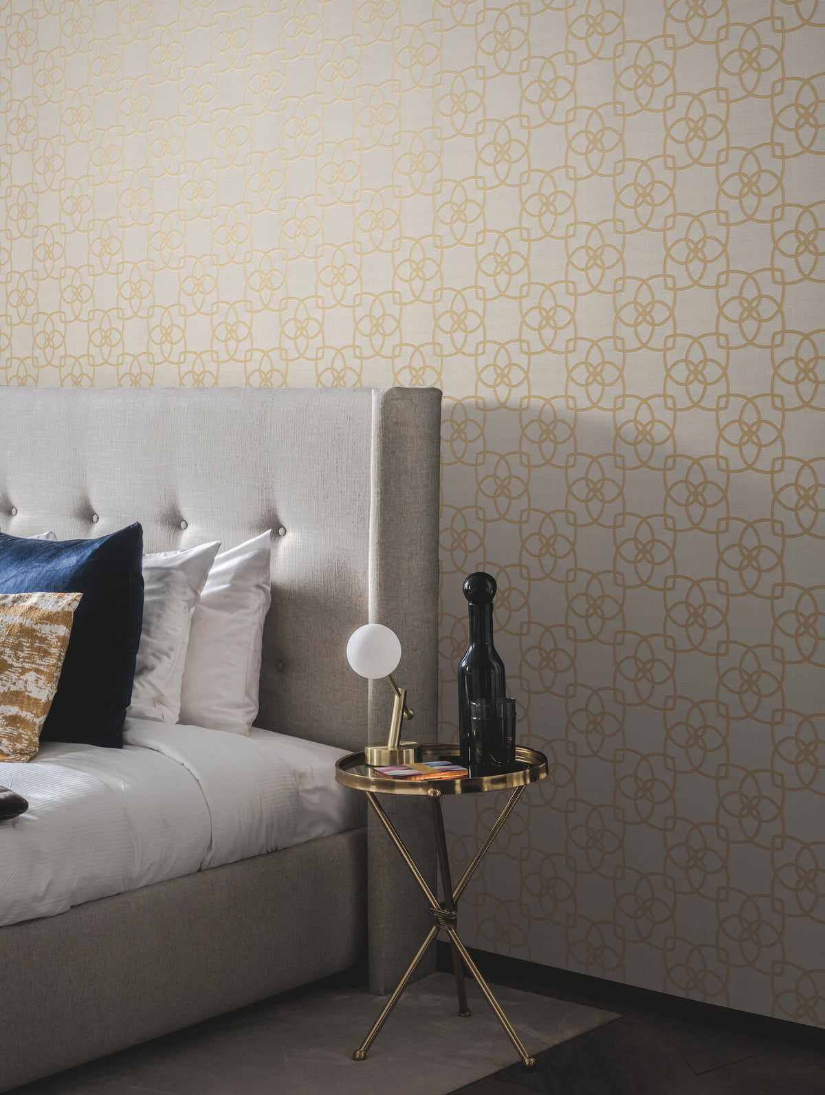 A modern bedroom featuring a beige tufted headboard, gold and blue pillows, and a round gold side table adorned with a lamp, books, and a dark bottle, set against York Wallcoverings’ Serendipity Wallpaper with elegant gold geometric patterns.