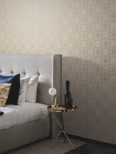 A modern bedroom featuring a gray upholstered bed with patterned pillows, a round gold side table displaying a lamp, black bottles, and magazines, set against a beige wall adorned with the Serendipity Wallpaper by York Wallcoverings in a gold geometric pattern.