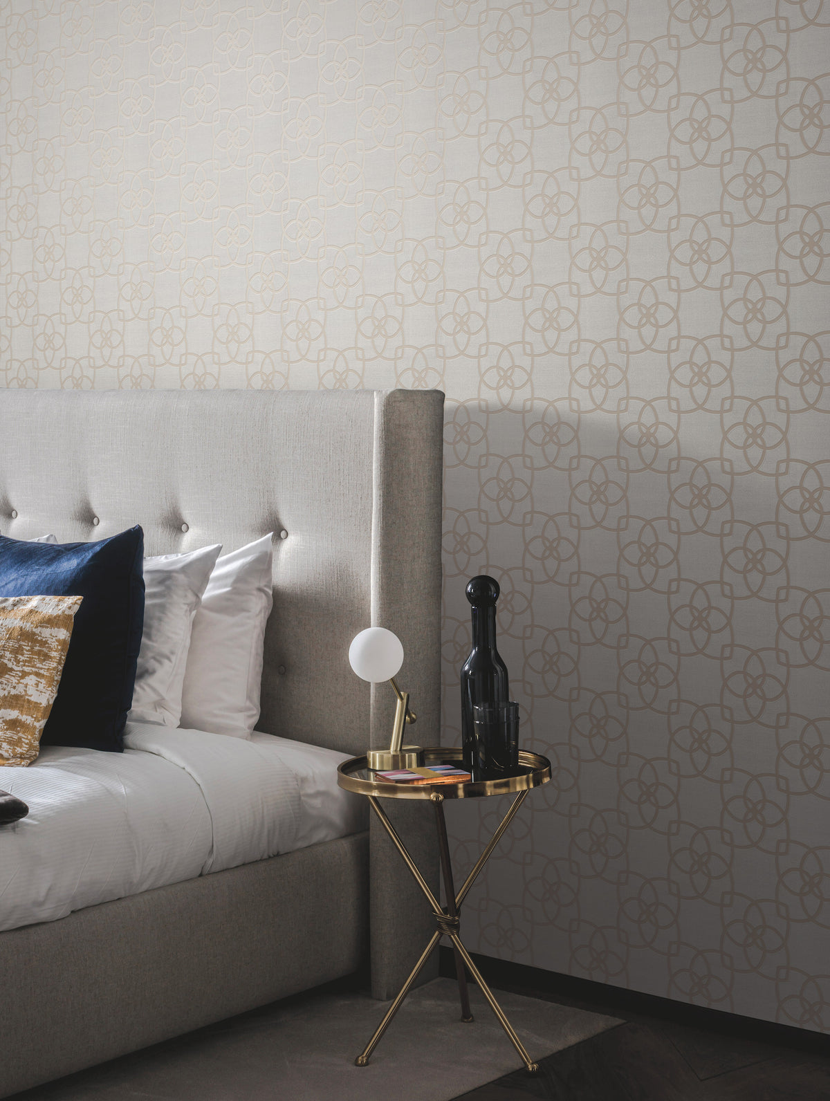 A modern bedroom featuring a gray upholstered bed adorned with blue and gold pillows, a round gold side table displaying a lamp, black bottle, and book, set against York Wallcoverings’ Serendipity Wallpaper in beige.
