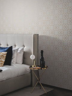 A modern bedroom featuring a gray upholstered bed, blue and gold pillows, and a small round gold side table topped with a lamp, a dark bottle, and a book. The wall is adorned with York Wallcoverings’ Serendipity Wallpaper, showcasing a subtle geometric pattern.