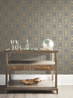 A modern bar cart with glassware, a bottle, a silver decorative orb, and a piece of driftwood sits against a gray wall adorned with York Wallcoverings’ Serendipity Wallpaper in a gold geometric pattern.