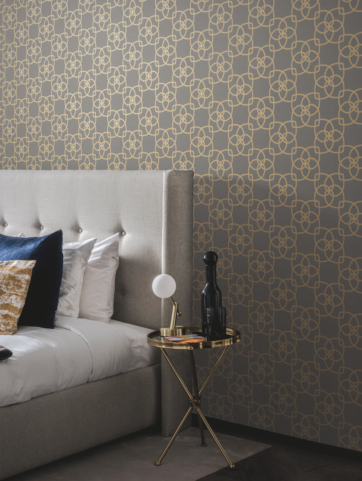 A modern bedroom featuring a tufted gray bed with white and blue pillows, a small round gold side table topped with a lamp, black bottle, and book, is set against a gray wall adorned with York Wallcoverings’ Serendipity Wallpaper in a gold geometric pattern.