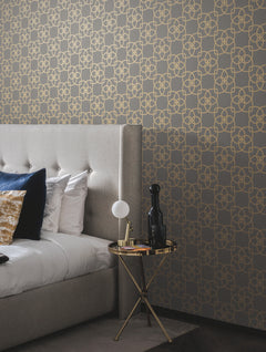 A modern bedroom featuring a tufted gray bed with white and blue pillows, a small round gold side table topped with a lamp, black bottle, and book, is set against a gray wall adorned with York Wallcoverings’ Serendipity Wallpaper in a gold geometric pattern.