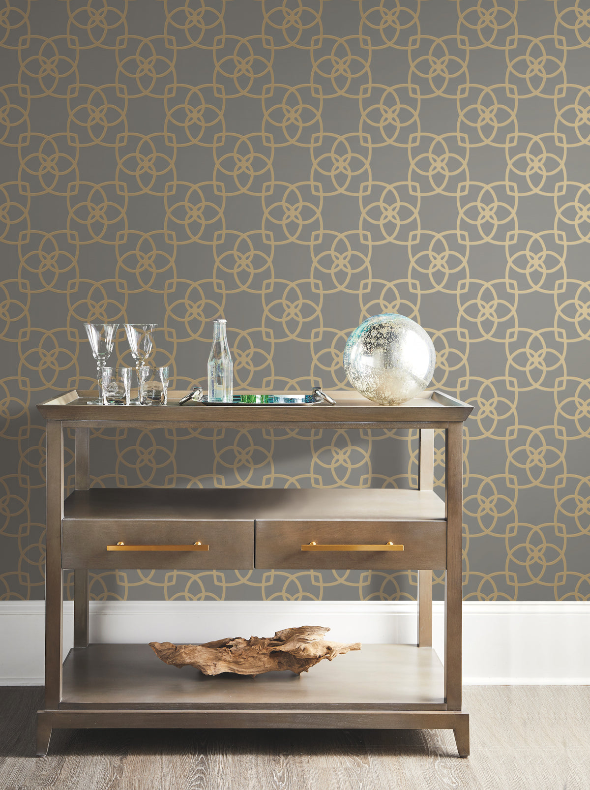 A small wooden bar cart with glassware, a bottle, and a decorative glass globe is set against the Serendipity Wallpaper by York Wallcoverings, featuring a gold geometric pattern. A large piece of driftwood rests on the bottom shelf.