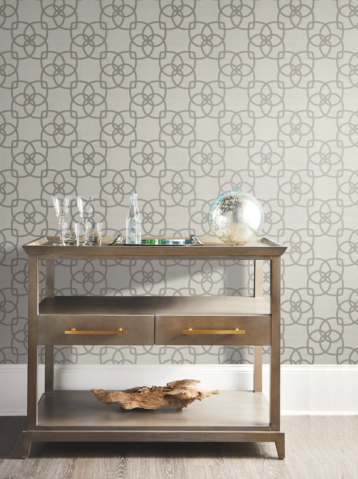 A wooden console table adorned with glassware, a glass bottle, a decorative glass sphere, and driftwood sits against a wall covered in York Wallcoverings’ Serendipity Wallpaper.