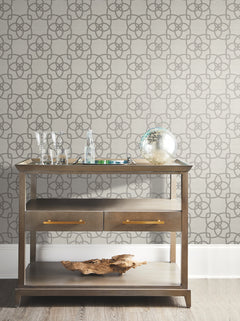 A wooden console table adorned with glassware, a glass bottle, a decorative glass sphere, and driftwood sits against a wall covered in York Wallcoverings’ Serendipity Wallpaper.