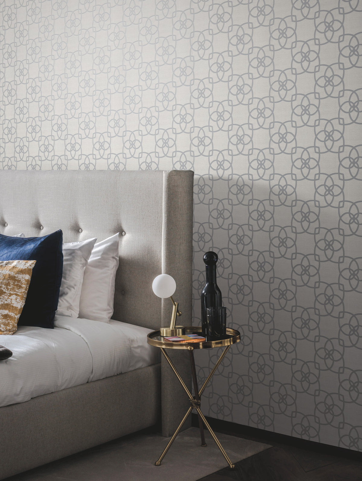 A modern bedroom featuring York Wallcoverings’ Serendipity Wallpaper, a tufted gray headboard, pillows, and a round gold side table displaying a lamp, a bottle, and a book.
