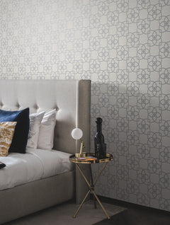 A modern bedroom featuring York Wallcoverings’ Serendipity Wallpaper, a tufted gray headboard, pillows, and a round gold side table displaying a lamp, a bottle, and a book.