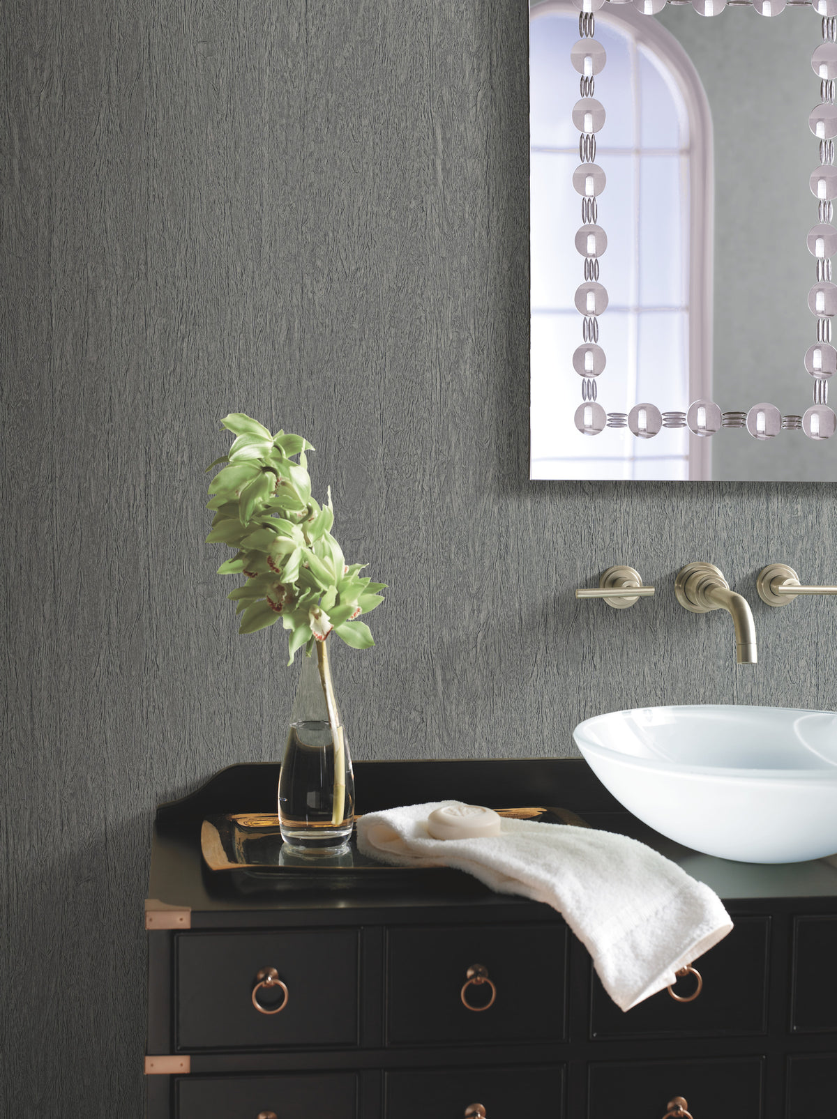 A modern bathroom featuring York Wallcoverings’ Natural Texture Wallpaper, paired with a black vanity cabinet with gold handles, a white bowl sink, wall-mounted faucet, a vase with green leaves, a folded towel, and a partial view of a mirror with a decorative border.