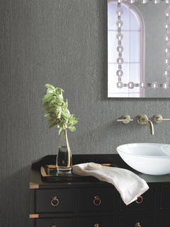 A modern bathroom featuring York Wallcoverings’ Natural Texture Wallpaper on the walls, complemented by a black vanity, vessel sink, wall-mounted faucet, towel, glass vase with green leaves, and a partially visible decorative mirror.