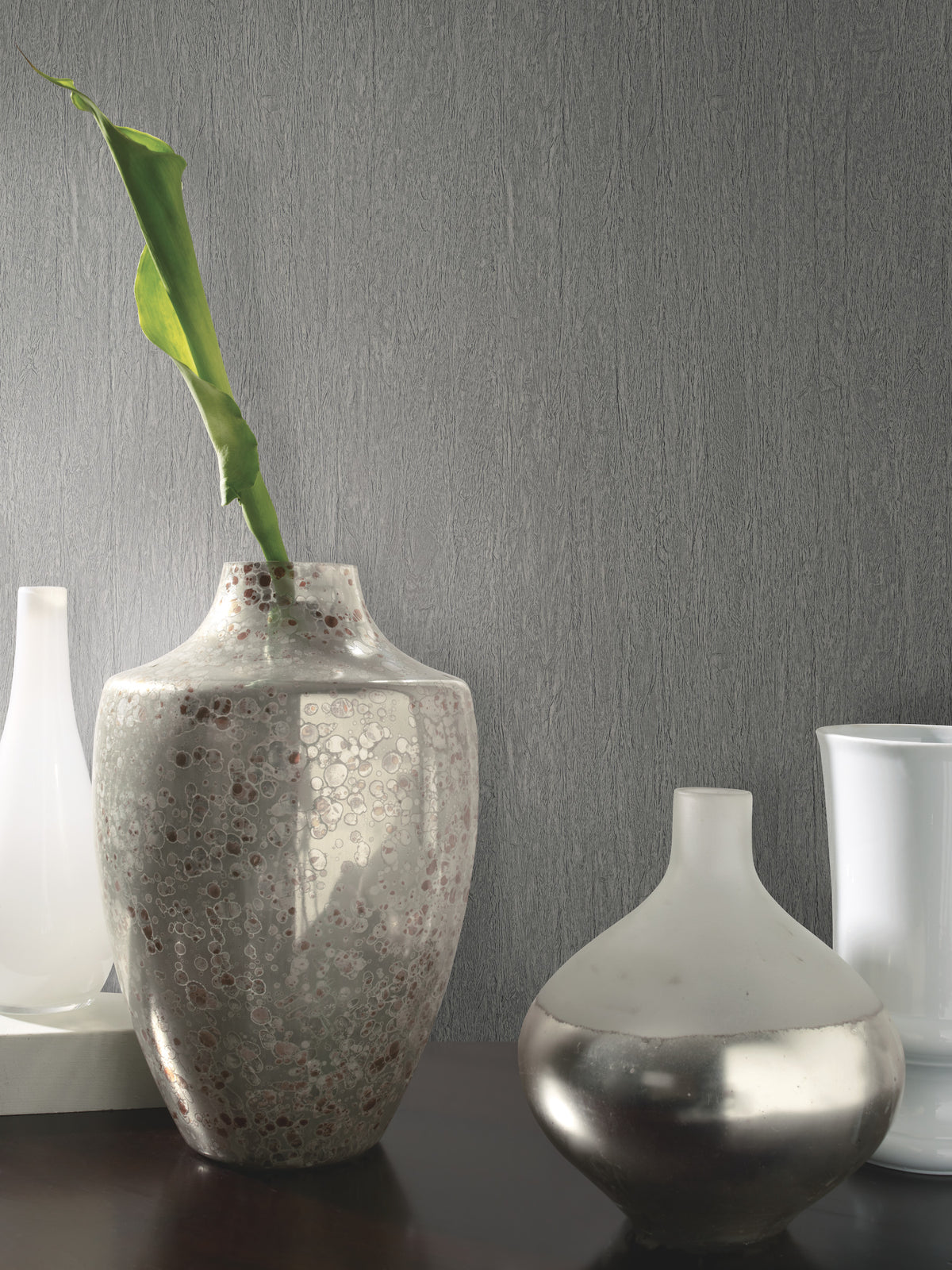 A textured gray vase with a single green leaf stands beside a small, round, frosted silver vase on a dark surface against a wall covered in York Wallcoverings’ Natural Texture Wallpaper. White vases are visible in the background.