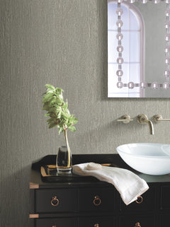 A modern bathroom features York Wallcoverings’ Natural Texture Wallpaper, a black vanity, a white vessel sink, a towel, and a vase with green leaves. Above is a decorative mirror reflecting a window with an arched top.