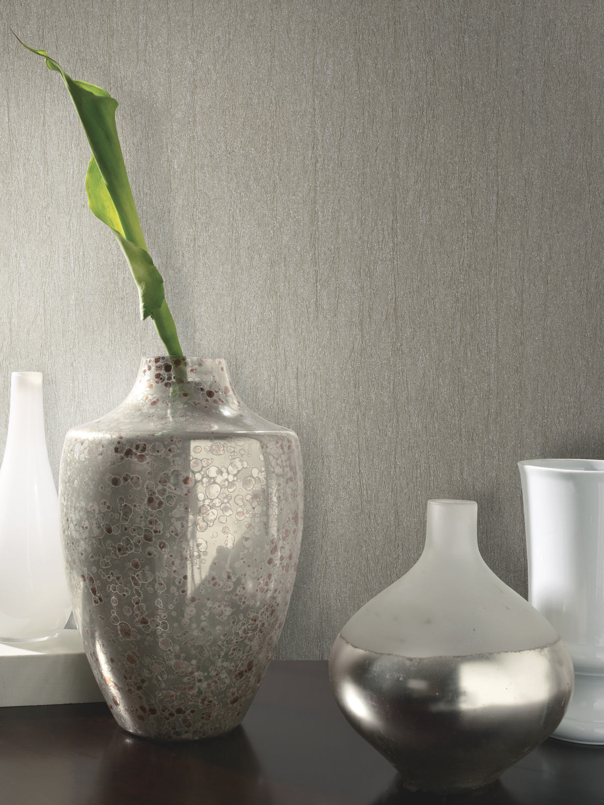 Three decorative vases sit on a dark surface against a wall adorned with Natural Texture Wallpaper by York Wallcoverings. The center vase is speckled with a single green leaf, the right vase is round and metallic, and the left vase is tall and white.