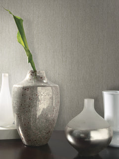 Three decorative vases sit on a dark surface against a wall adorned with Natural Texture Wallpaper by York Wallcoverings. The center vase is speckled with a single green leaf, the right vase is round and metallic, and the left vase is tall and white.