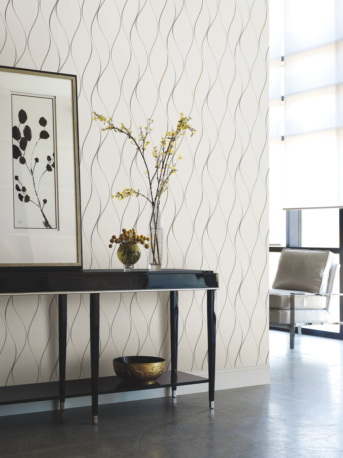 Modern interior featuring a black console table, framed botanical art, a vase with yellow flowers, a small decorative bowl, and a cushioned chair by large sunlit windows. The background showcases York Wallcoverings’ Wavy Stripe Wallpaper.