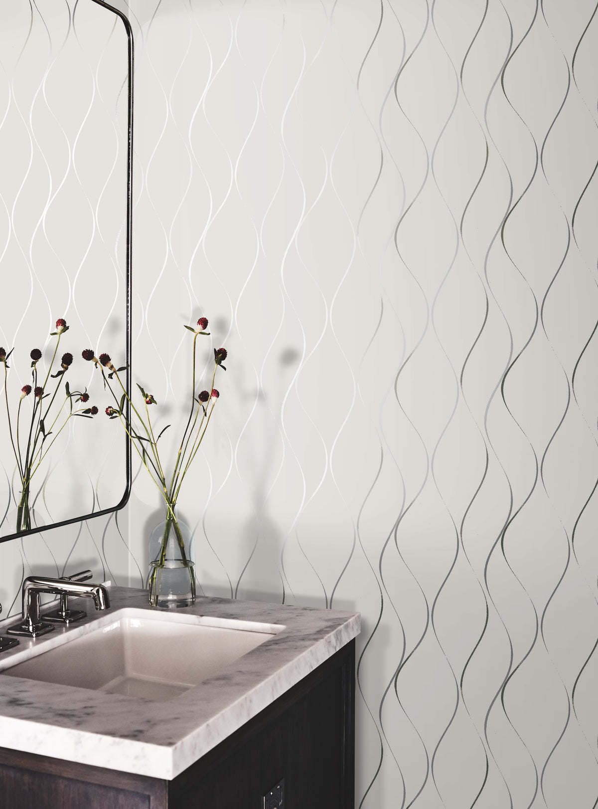 A bathroom vanity with a white sink, marble countertop, and a dark wood cabinet. A mirror and a glass vase with flowers sit on the counter. The wall features York Wallcoverings’ Wavy Stripe Wallpaper, adding a textured pattern to the space.