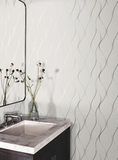 A bathroom vanity with a white sink, marble countertop, and a dark wood cabinet. A mirror and a glass vase with flowers sit on the counter. The wall features York Wallcoverings’ Wavy Stripe Wallpaper, adding a textured pattern to the space.