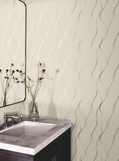 A modern bathroom corner featuring a marble sink, a rectangular mirror, and a glass vase with long-stemmed flowers. The wall is adorned with Wavy Stripe Wallpaper by York Wallcoverings, showcasing a beige background accentuated by elegant wavy silver lines.