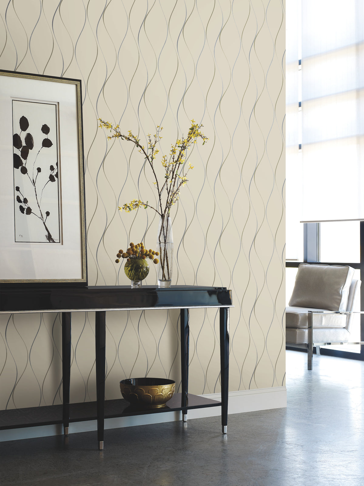 A modern hallway features a black console table, a vase of yellow flowers, framed art, a ceramic bowl, and a chair by tall windows. The wall is adorned with York Wallcoverings’ Wavy Stripe Wallpaper in beige.