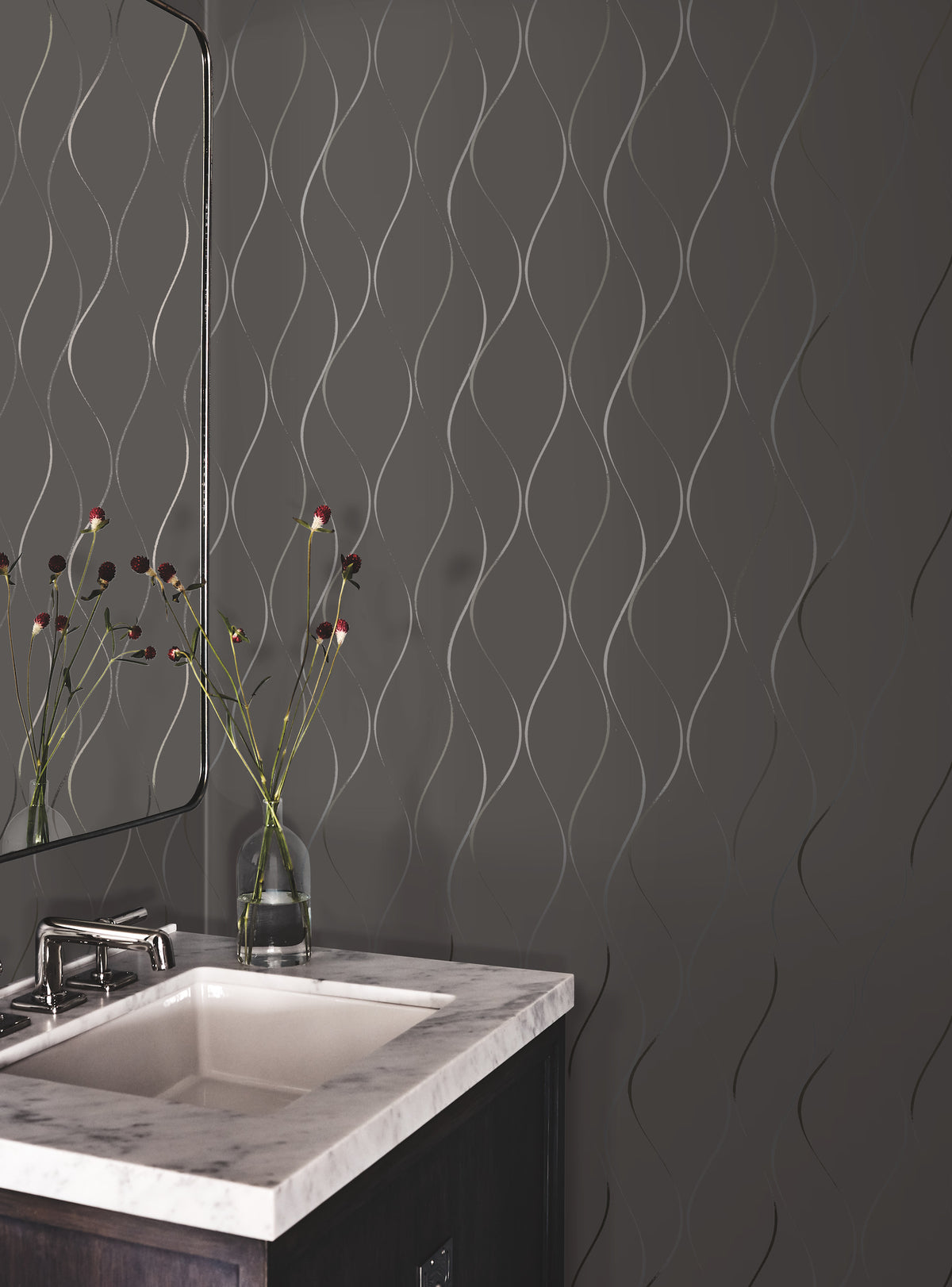 A modern bathroom featuring the Wavy Stripe Wallpaper by York Wallcoverings on a gray wall, complemented by a marble sink, a mirror, and a vase with long-stemmed flowers.