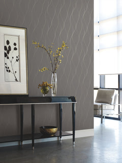 Modern interior featuring a black console table, framed botanical artwork, a vase with yellow flowers, a decorative bowl, and a beige armchair by large windows. The wall is adorned with York Wallcoverings’ Wavy Stripe Wallpaper for a subtle wavy pattern.