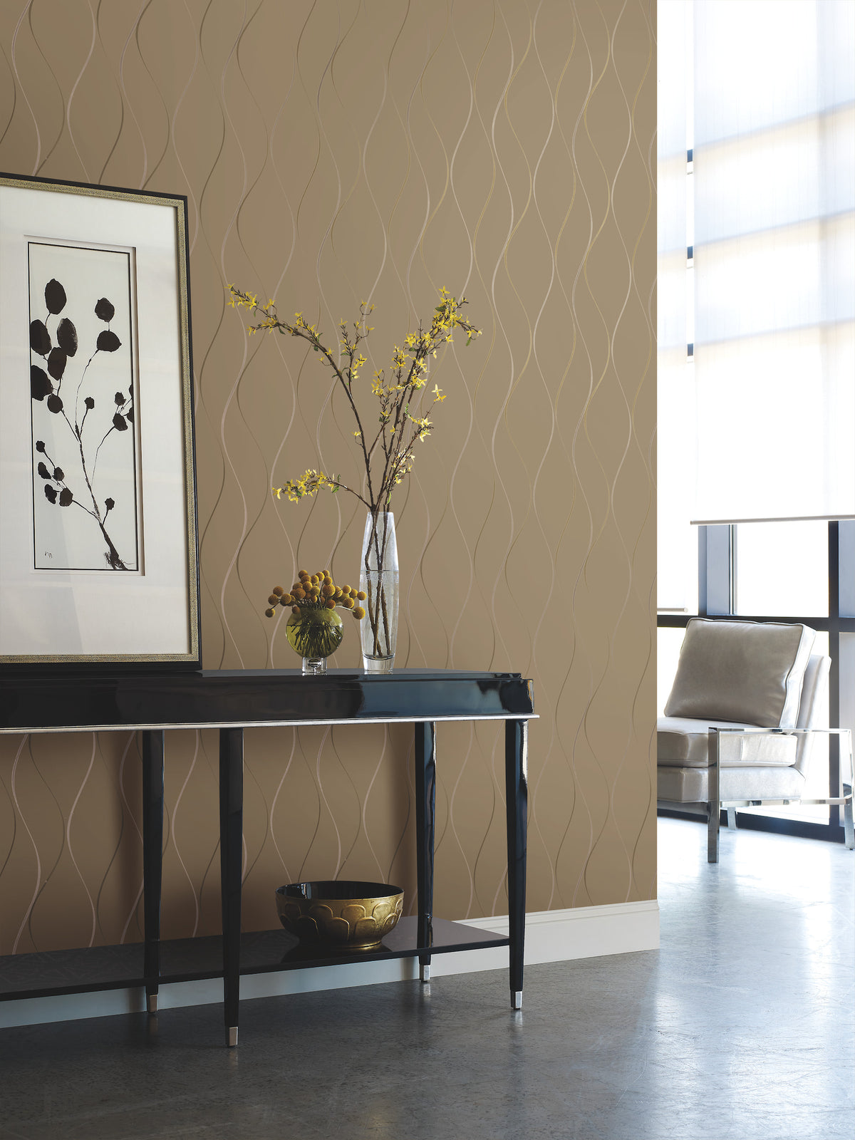 A modern interior featuring York Wallcoverings’ Wavy Stripe Wallpaper in a tan pattern, a black console table adorned with a vase of yellow flowers, framed botanical art, a decorative bowl, and a metallic chair positioned by large windows with sheer curtains.